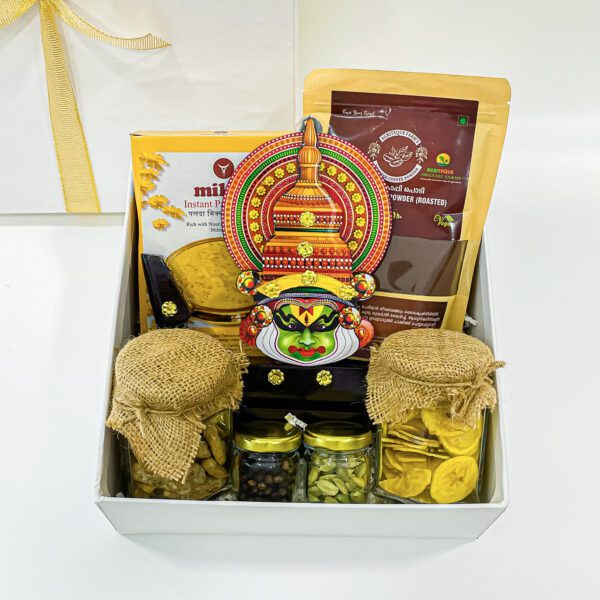 An assortment of traditional Kerala-style handicraft gift items, including a Kathakali head boat, Organic coffee powder 250g, an Instant payasam mix 200g, a Banana chips bottle, Japggery chips bottle, and Black pepper, Cardamom displayed in a gift box